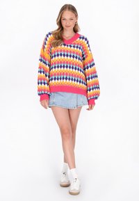 Pulover colorat tricotat cu un model în diamant în nuanțe de roz, mov, galben și turcoaz; croială lejeră cu decolteu în V și manșete dantelate, asortat cu o fustă din denim.