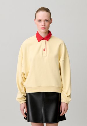 Femme aux cheveux plaqués en arrière portant un sweat-shirt jaune pâle avec un col rouge et une jupe en cuir noire sur un fond gris clair uni.