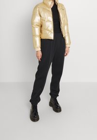 Złota kurtka puchowa o błyszczącej powierzchni, z zamek błyskawicznym i stójką, w zestawie z czarnymi dresami i masywnymi czarnymi butami.