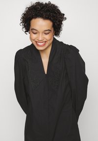 Blusa preta com um colarinho largo apresentando detalhes florais bordados. Mangas com franzido e fecho de botão na frente. Textura suave.