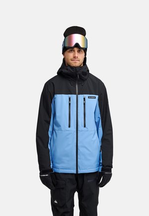 Man draagt een zwarte en blauwe winterjas, zwarte handschoenen, zwarte broek, zwarte muts en reflecterende skibril op het voorhoofd.