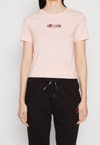 Kobieta w jasnoróżowej koszulce Armani Exchange i czarnych spodniach z troczkami, z logo Armani na klatce piersiowej i udzie.