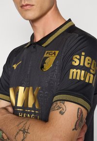 Crna sportska majica s zlatnim detaljima, s uzorkom na tkanini, ovratnikom, logotipom i podebljanim tekstom na rukavima.