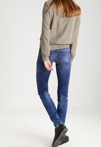 Femme debout, jambes croisées, portant une veste beige, un jean bleu et des bottines noires sur un fond blanc uni.