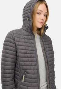 Grijze pufferjack met horizontale naden, een strakke capuchon, ritszakken en een gladde afwerking, gedragen over een lichtgrijze top.