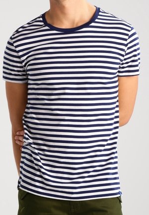 Homme portant un T-shirt à manches courtes rayé horizontalement bleu marine et blanc, debout avec un bras derrière le dos.