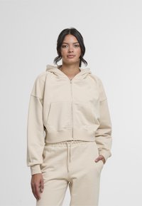 Bež zip-up hoodie napravljen od mekog materijala, sa velikom kapuljačom, prednjim džepom i rebrastim manžetama, u kombinaciji sa odgovarajućim hlačama.