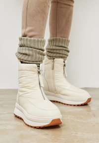 Botine albe umflate la glezne, cu fermoare pe lateral și mansete din tricot texturat, având o talpă groasă din cauciuc de o culoare brună contrastantă.