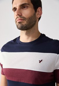 Navy, white, and burgundy striped t-shirt with cotton fabric. Features a small embroidered logo on the chest. Round neckline and short sleeves.