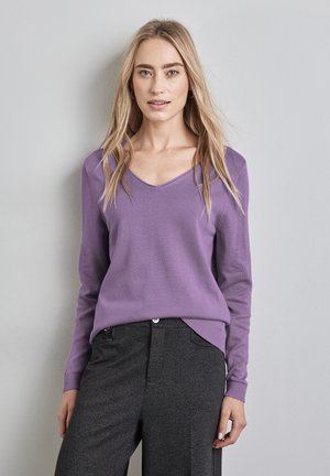 Lila gestrickter Pullover mit V-Ausschnitt, langen Ärmeln und gerippten Akzenten, kombiniert mit hochgeschnittenen dunkelgrauen Hosen.