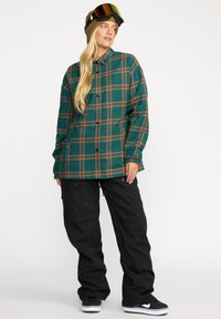 Veste oversize à carreaux verts et bruns avec fermeture à boutons, associée à un pantalon cargo noir et des baskets noires avec des semelles blanches.