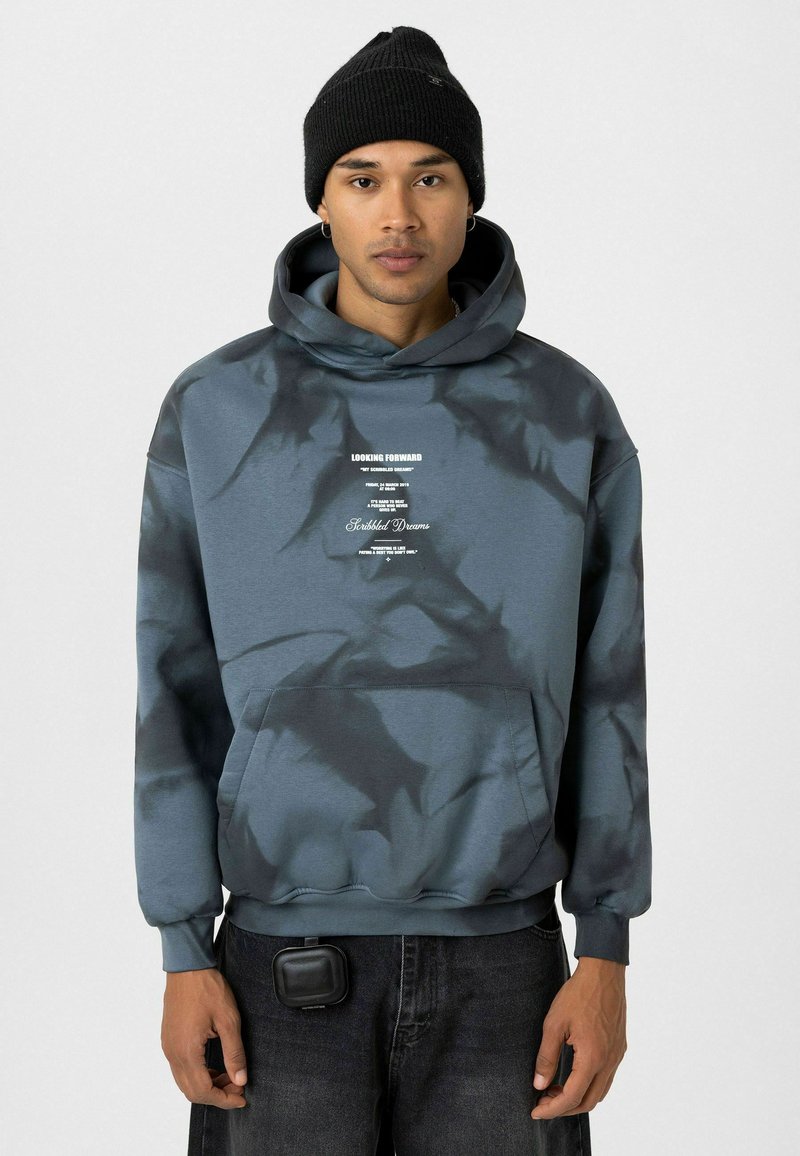 Hoodie in blauwgrijs met een tie-dye patroon, voorzien van een voork pocket en geprinte branding. Gecombineerd met donkere jeans en een zwarte beanie.