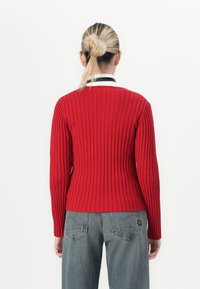 Blonde vrouw met een bun draagt een rode geribde trui en grijze jeans, terwijl ze met haar rug naar de camera staat op een witte achtergrond.