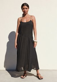 Robe longue noire à fines bretelles, coupe décontractée et tissu texturé. Accessoirisée d'un bracelet épais et portée avec des sandales plates.