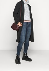 Schwarzer langer Cardigan, grauer gestrickter Rollkragenpullover, hellrosa mit gewelltem Saum, blaue Slim-Fit-Jeans und schwarze schnürbare Kampfstiefel. Burgunderfarbene Umhängetasche.