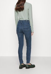 Jeans ajustados de denim azul oscuro con cintura alta, que cuentan con dos bolsillos traseros y un acabado de textura suave. Combinados con una blusa verde de manga larga.