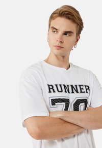 Hvid kortærmet t-shirt med sort tekst "RUNNER 70" med et prikket mønster. Rund hals, letvægtsstof, afslappet pasform.