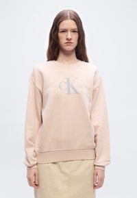 Γυναίκα με μακριά καστανά μαλλιά που φορ άει ανοιχτό ροζ φούτερ Calvin Klein και μπεζ φούστα, στέκεται μπροστά σε απλό λευκό φόντο.