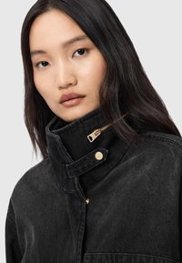 Veste en denim noire avec un col montant, fermeture éclair dorée et accents en boutons. Le tissu semble texturé avec des détails de couture visibles.
