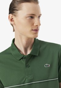 Polo verde hecho de tela texturizada, con cuello, tres botones y una franja blanca en el pecho. Logotipo de Lacoste en el lado izquierdo.