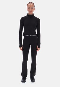 Juodas sportinis kombinezonas su aukšta apykakle, pilnais užtrauktukais ir prigludusia forma. Yra žymėta juosmens juosta ir platėjančios kelnės.