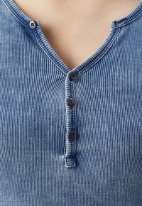 Blå ribbad henley-tröja med V-urringning, fem svarta knappar och ett strukturerat tyg. Designen inkluderar subtila vertikala randiga mönster.