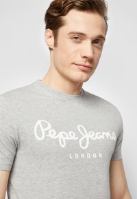 Pelēks kokvilnas T-krekls ar baltu kursīvu "Pepe Jeans" logo un "LONDON" tekstu, īsām piedurknēm un apaļu kakla izgriezumu.