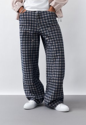 Άτομο που φορά χαλαρά, μπλε και γκρι καρό παντελόνια με πλαϊνές τσέπες, λευκά αθλητικά παπούτσια και ελαφρύ μπουφάν, με τα χέρια στις τσέπες.