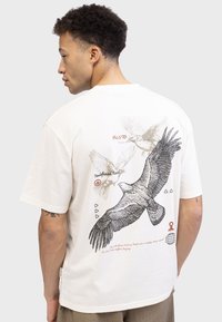 T-shirt en coton crème avec un grand motif d'oiseau noir au dos, accentué par un texte rouge et des formes géométriques. Manches courtes, coupe décontractée.