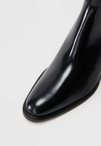 Bota de cuero negro brillante con punta cuadrada y tacón bajo, mostrada contra un fondo blanco liso.