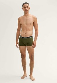Boxers verts olive avec une ceinture élastique blanche portant la marque. Texture lisse et coupe ajustée, présentés sur un modèle masculin torse nu.
