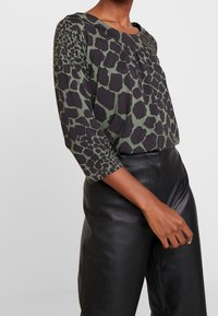 Zwarte en groene gebloemde blouse met een ronde halslijn en driekwart mouwen, gecombineerd met zwarte leren broek, met nadruk op texturen.