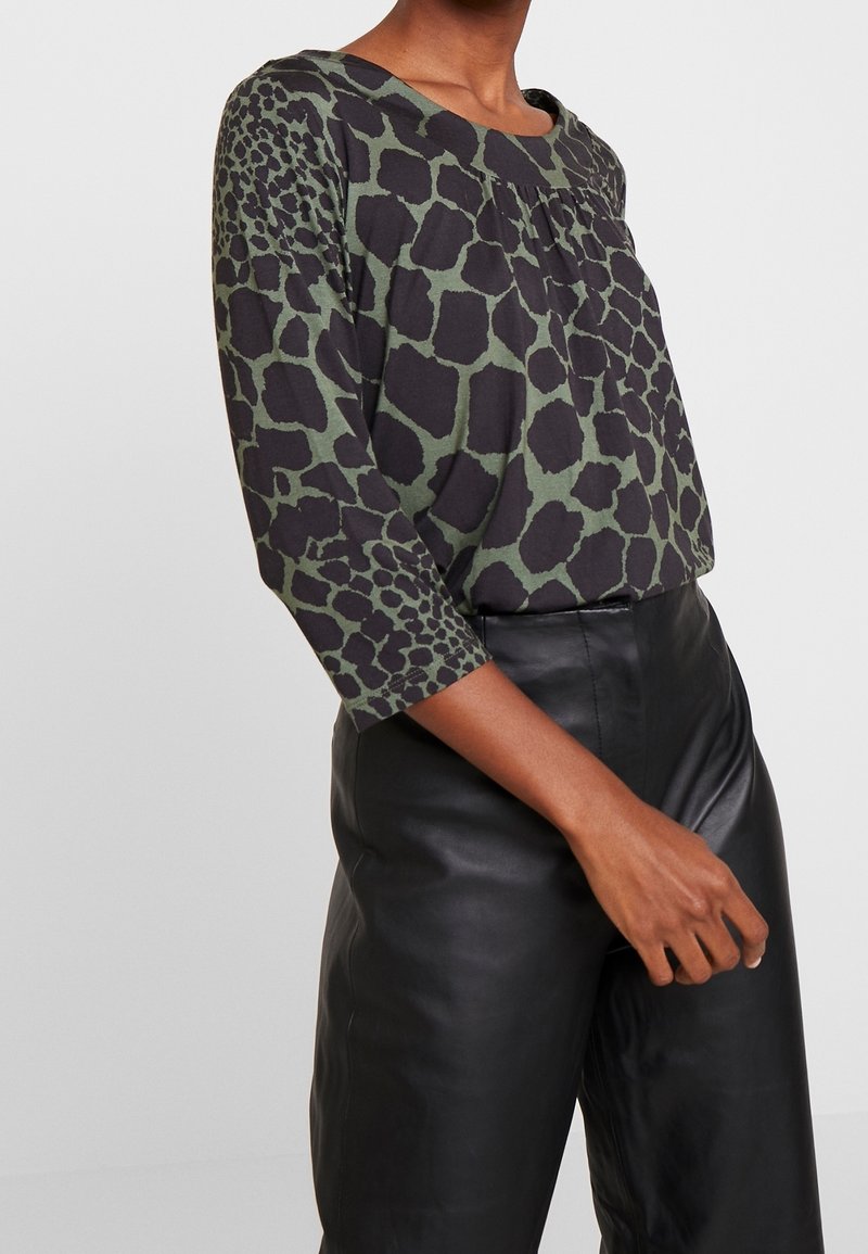 Zwarte en groene gebloemde blouse met een ronde halslijn en driekwart mouwen, gecombineerd met zwarte leren broek, met nadruk op texturen.
