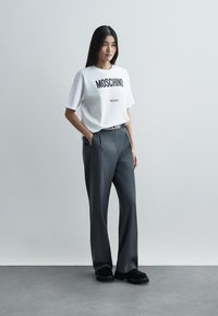 Hvid grafisk t-shirt med teksten "Moschino Milano", parret med grå brede bukser med en logo-elastik i taljen og sorte sko.