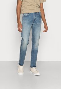 Ljust blå denimjeans med rak passform, blekt mönster och femfickdesign, kombinerat med beige sneakers och en tan t-shirt.
