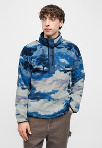 Νέος άνδρας που φοράει μπλε και γκρι φλις πουλόβερ με μοτίβο, φερμουάρ μπροστά και καφέ παντελόνι, στέκεται μπροστά σε λευκό απλό φόντο.