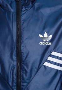 Tummansininen tuuletinjakku, jossa on vetoketjusuljenta, herringbone-kuviointi, valkoiset kolme viivaa ja Adidas-logo.