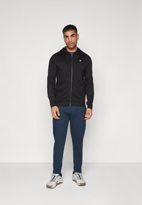 Felpa nera con zip, cappuccio e logo giallo, abbinata a pantaloni blu scuro e sneaker grigie e nere. Sfondo semplice.
