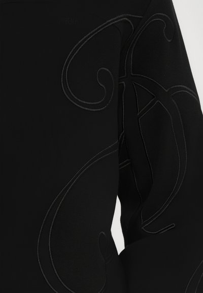 Vêtement noir avec une broderie en relief complexe dans un design de fil contrasté, mettant en valeur des formes courbes sur la manche.