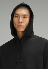 Sudadera negra con una capucha grande, mangas raglán y cordones. Textura suave, diseño minimalista y sin patrones ni logotipos visibles.