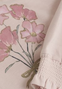 Ljust rosa tyg med ett blommönster och glittriga detaljer. Överdelen har en rynkad manschett med en volangkant och en dekorativ rosett.