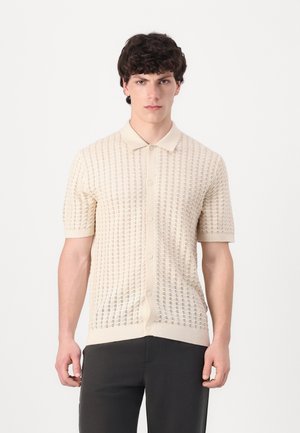 Short-sleeve, cream-colored knit polo shirt with a textured open stitch pattern and a button-down front. Features a classic collar and ribbed cuffs.