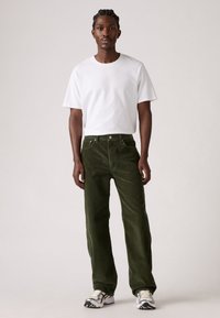 Man in een witte korte mouw t-shirt, donkergroene corduroy broek en witte sneakers, tegen een effen lichte achtergrond.
