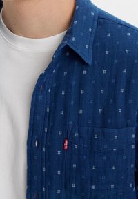 Camicia blu a fantasia realizzata in denim, con design button-up, una tasca a toppa e una piccola etichetta rossa. La texture è intrecciata con accenti chiari.
