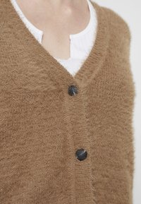 Brun stickad cardigan med en mjuk, fluffig textur, med en V-ringning och tre svarta knappar. Lager ovanpå en vit texturerad topp.