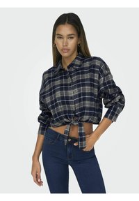 Camicia di flanella a quadri blu navy con davanti annodato, chiusura a bottoni e maniche lunghe. Abbinata a jeans in denim scuro.