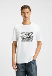 Witte T-shirt met korte mouwen met een zwart grafisch ontwerp met bloemaccenten en het woord "HUGO." Casual pasvorm. Draagt met blauwe denimjeans.