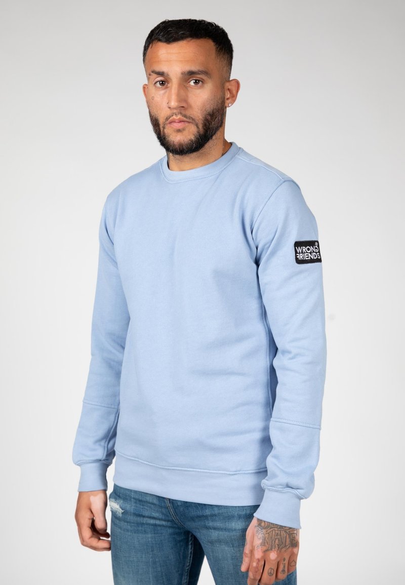 Sudadera azul claro de mezcla de algodón, con cuello redondo, puños acanalados y un parche negro con el logo "WRONG FRIENDS" en la manga.