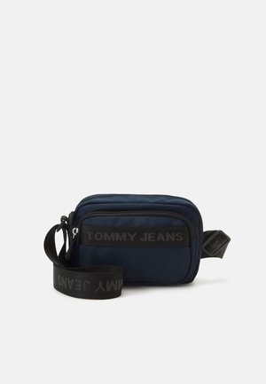 Granatowa torba na ramię Tommy Jeans z czarnym regulowanym paskiem i widoczną przednią kieszenią na zamek na jednolitym tle.