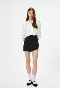 Camicia bianca con parte frontale a volant, mini gonna nera e ballerine nere. La modella indossa calze bianche fino al ginocchio e sta in piedi con le mani in tasca.